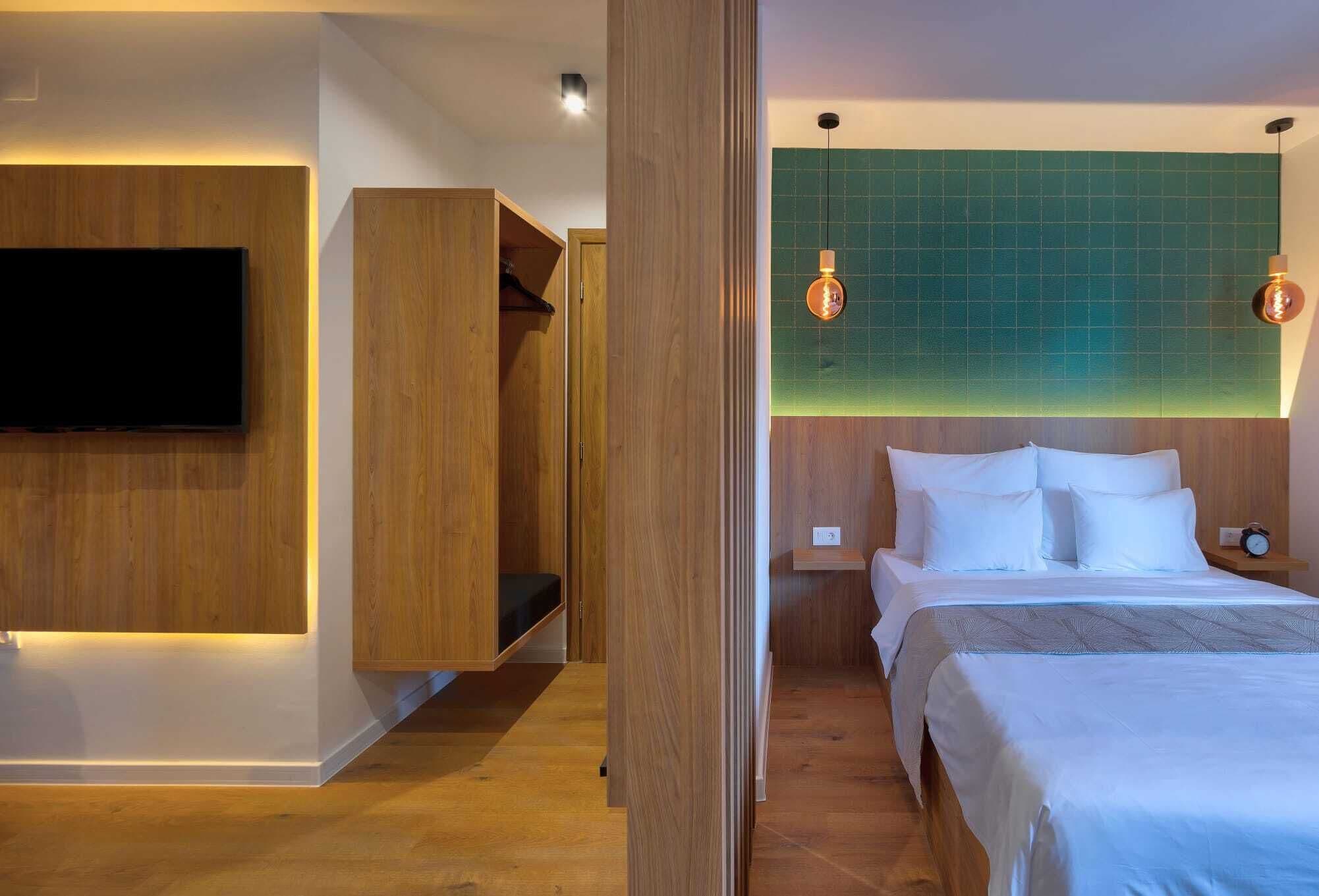 A modern hotel room with a large bed, a TV mounted on the wall, wooden furniture, and a green-tiled accent wall.