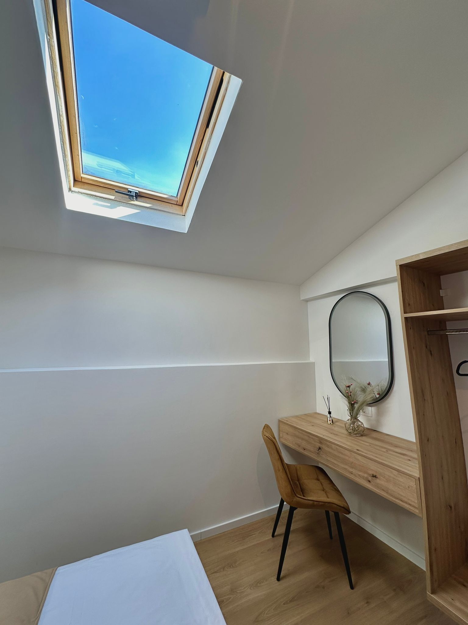 A bedroom with a skylight, a wooden desk, a chair, a bed, and a mirror on the wall.