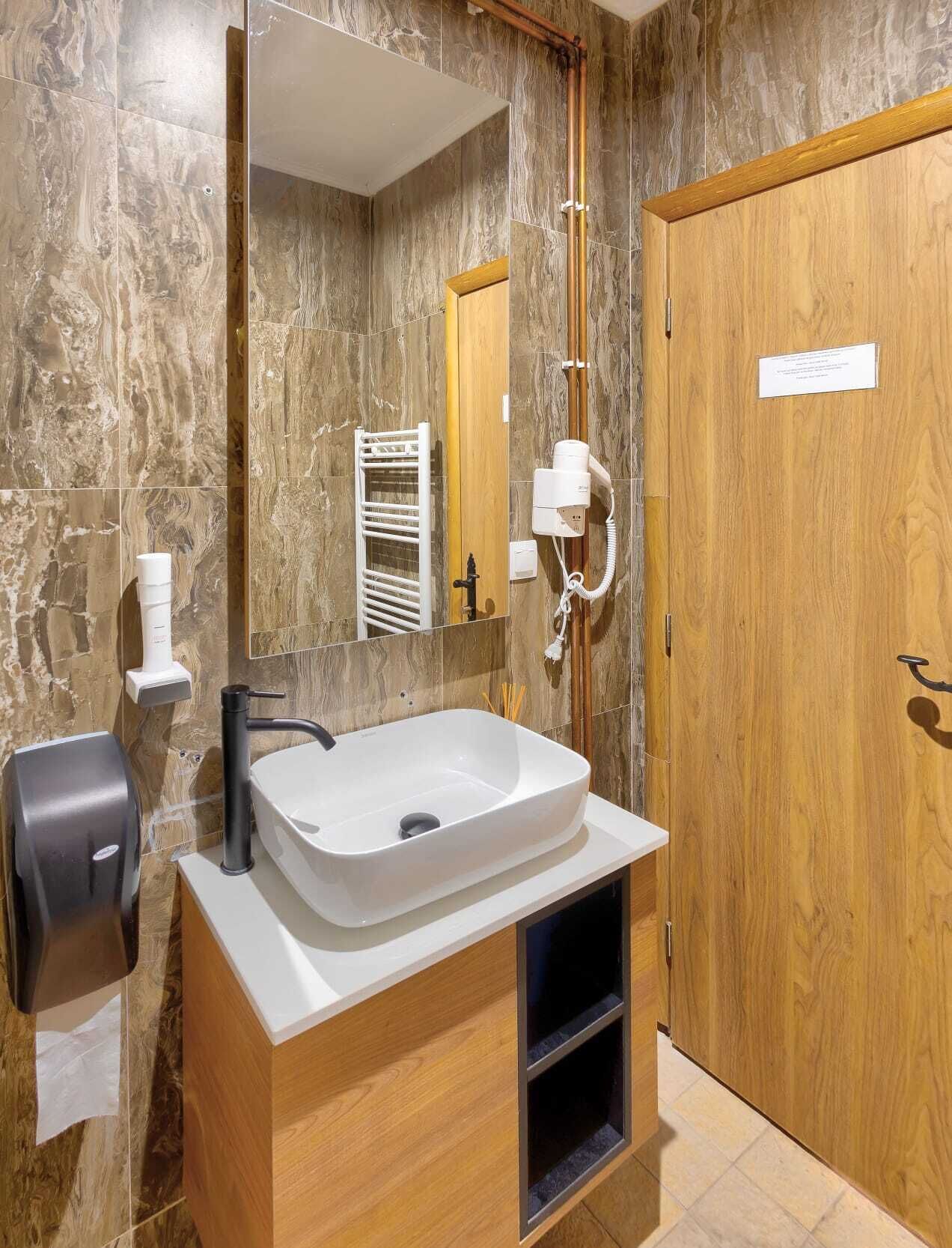 A modern bathroom with a marble wall, a large mirror, a white sink, a towel dispenser, and a wooden door.