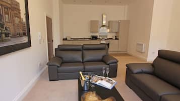 A modern living room with a black leather sofa, a wooden coffee table with a bottle and glasses, and a magazine. A kitchen with cabinets and appliances is visible in the background.