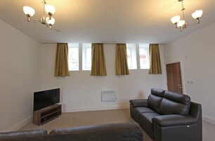 Una sala de estar con televisión de pantalla plana, un sofá de cuero y un sillón reclinable. Hay cuatro ventanas con cortinas, una lámpara de techo y una puerta.