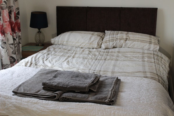 A neatly made bed with a folded towel, a dark headboard, and a lamp on a bedside table.