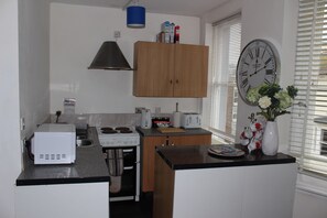 A compact kitchen with a microwave, stove, and toaster. There is a clock on the wall and a window with blinds.