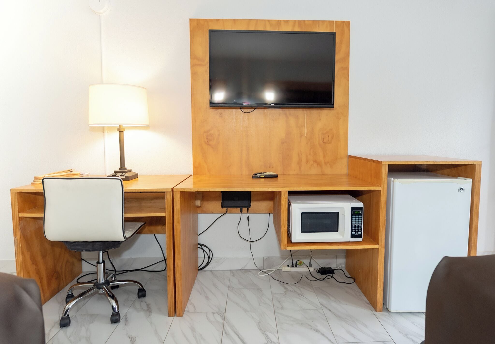 A compact hotel room with a wooden desk, a television mounted above it, a white chair, a lamp, a microwave, and a small refrigerator.