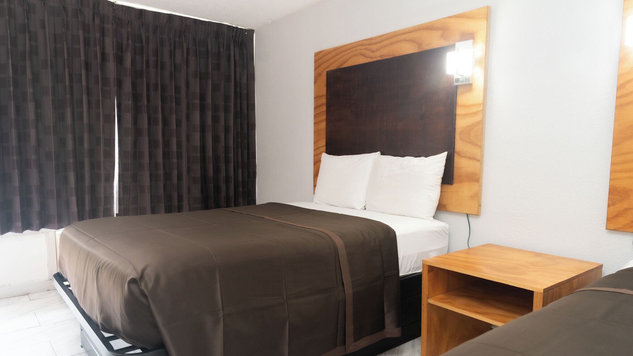 Habitación de hotel con cama, mesita de noche, cabecera de madera y ventana con cortinas.