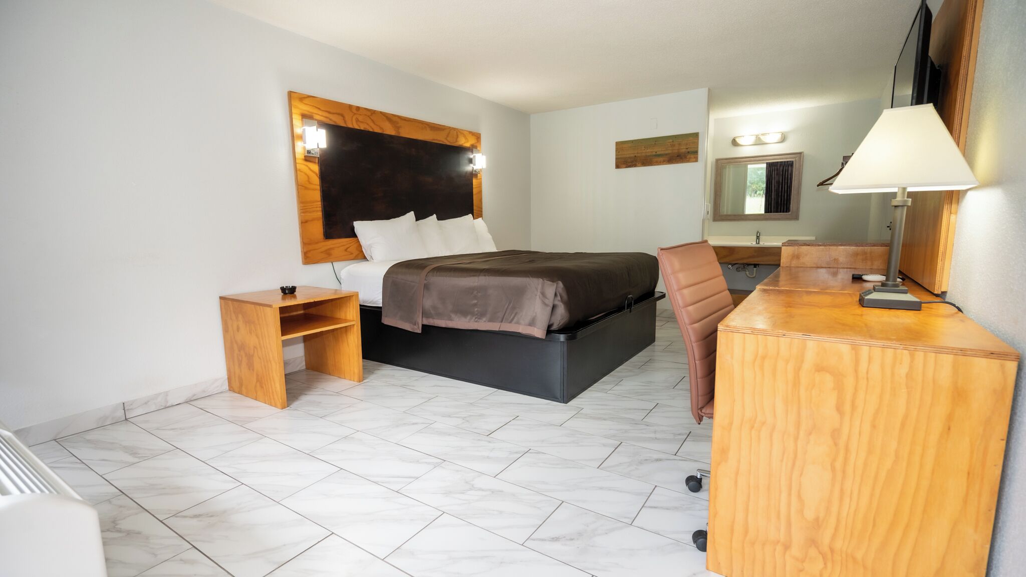 A hotel room with a bed, bedside table, chair, desk, and a mirror.