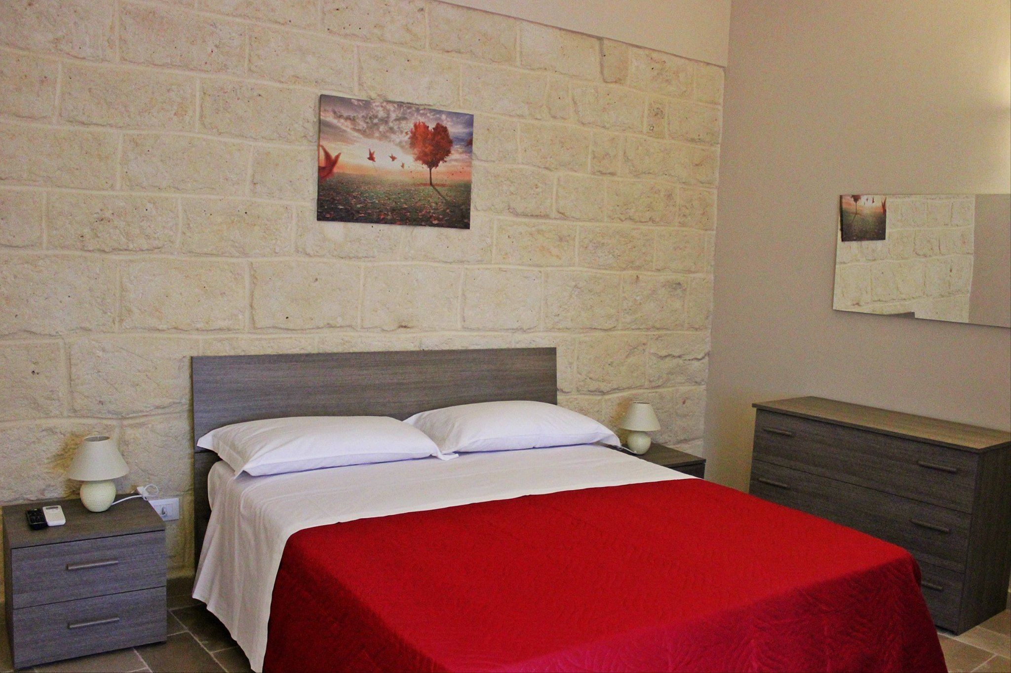 Una cama bien hecha con colcha roja, una mesita de noche con lámpara, un aparador y una pared de piedra con un cuadro.