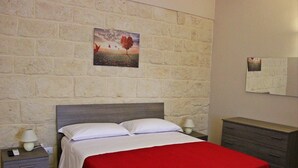 Una cama bien hecha con colcha roja, una mesita de noche con lámpara, un aparador y una pared de piedra con un cuadro.