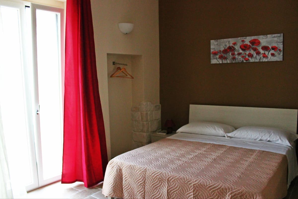 Ein Schlafzimmer mit einem Bett, einem roten Vorhang, einer Wandleuchte und einem Gemälde von roten Blumen.