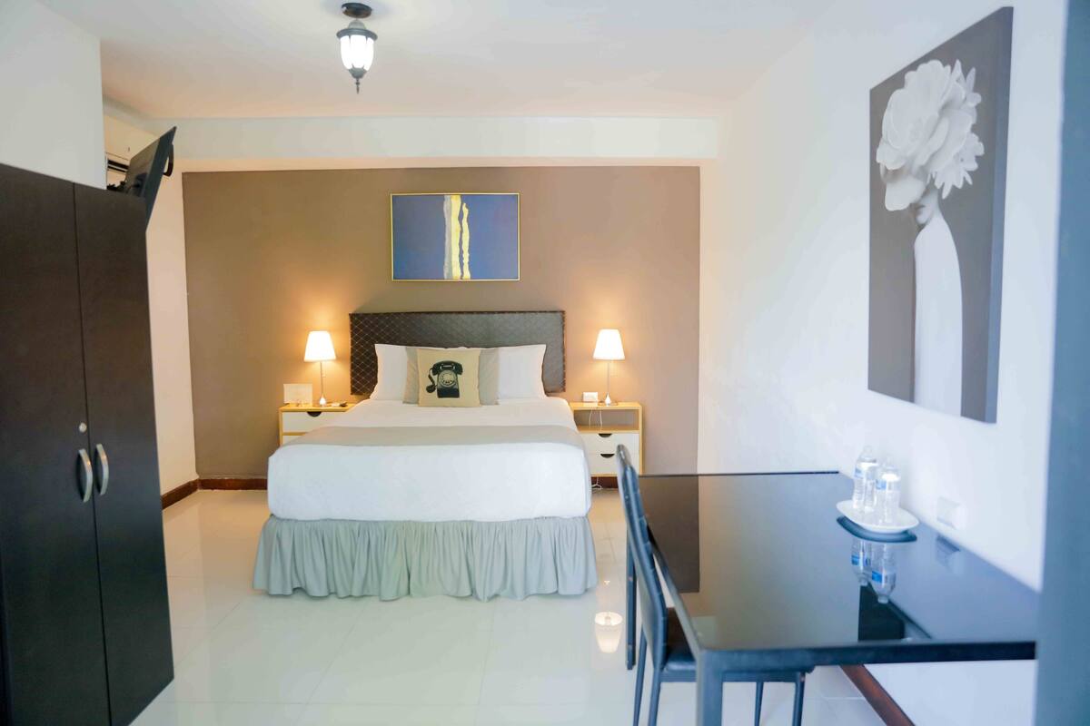 A hotel room with a large bed, two bedside tables with lamps, a desk with chairs, and a painting on the wall.