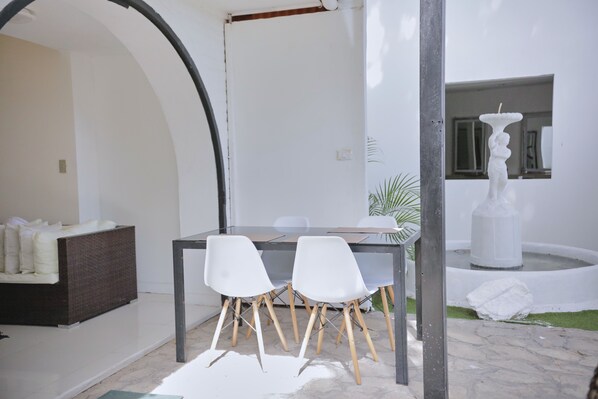 Un interior moderno con zona de comedor que incluye una mesa de vidrio y sillas blancas, un sofá blanco y una fuente.
