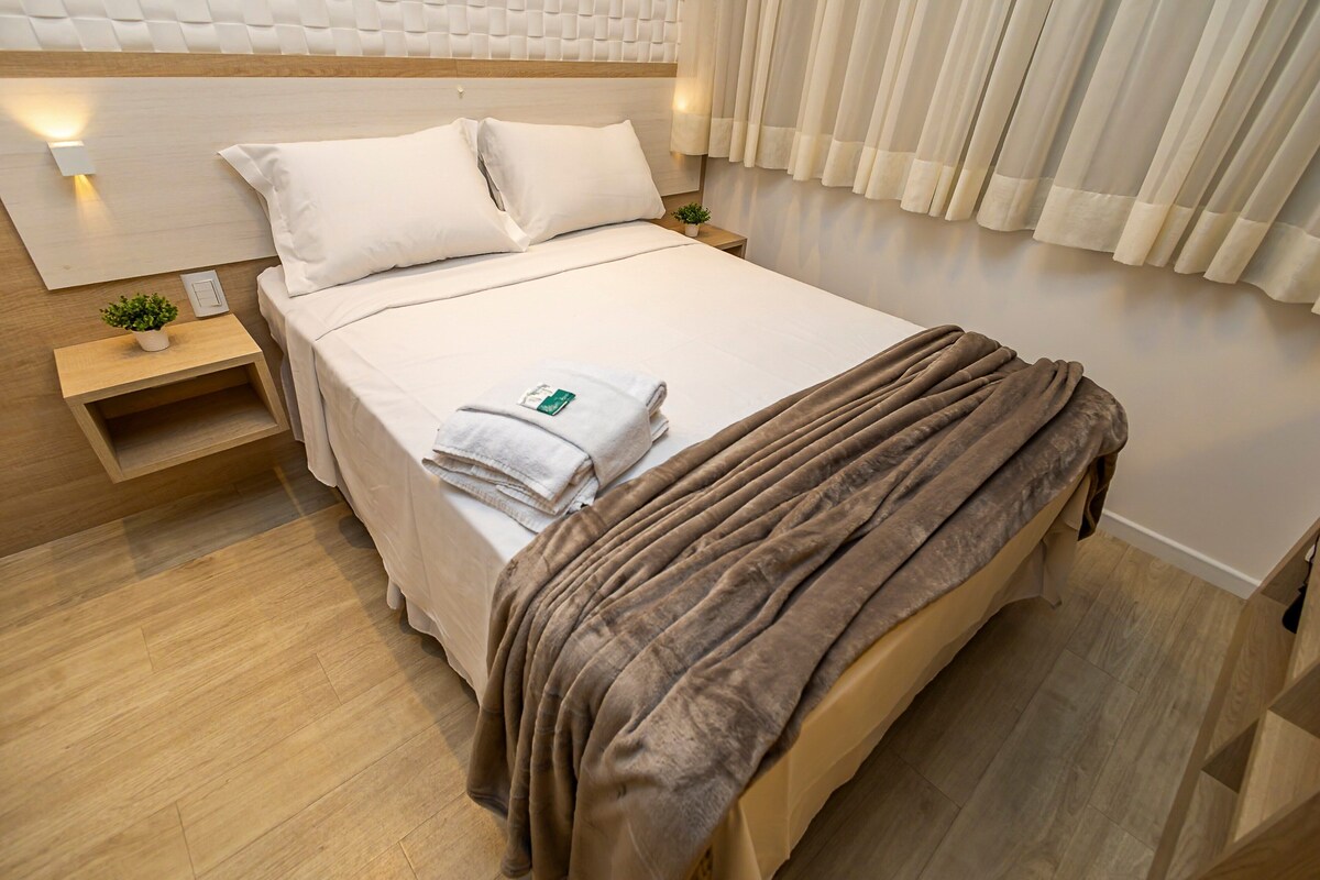 A modern hotel room with a wooden headboard, a neatly made bed with white linens, a brown throw blanket, and a small bedside table with a plant and a lamp.
