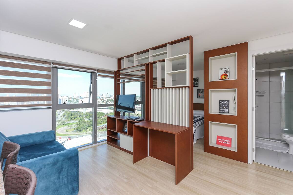 Quarto moderno com uma janela grande, um sofá azul, uma escrivaninha de madeira com um computador e um chuveiro.