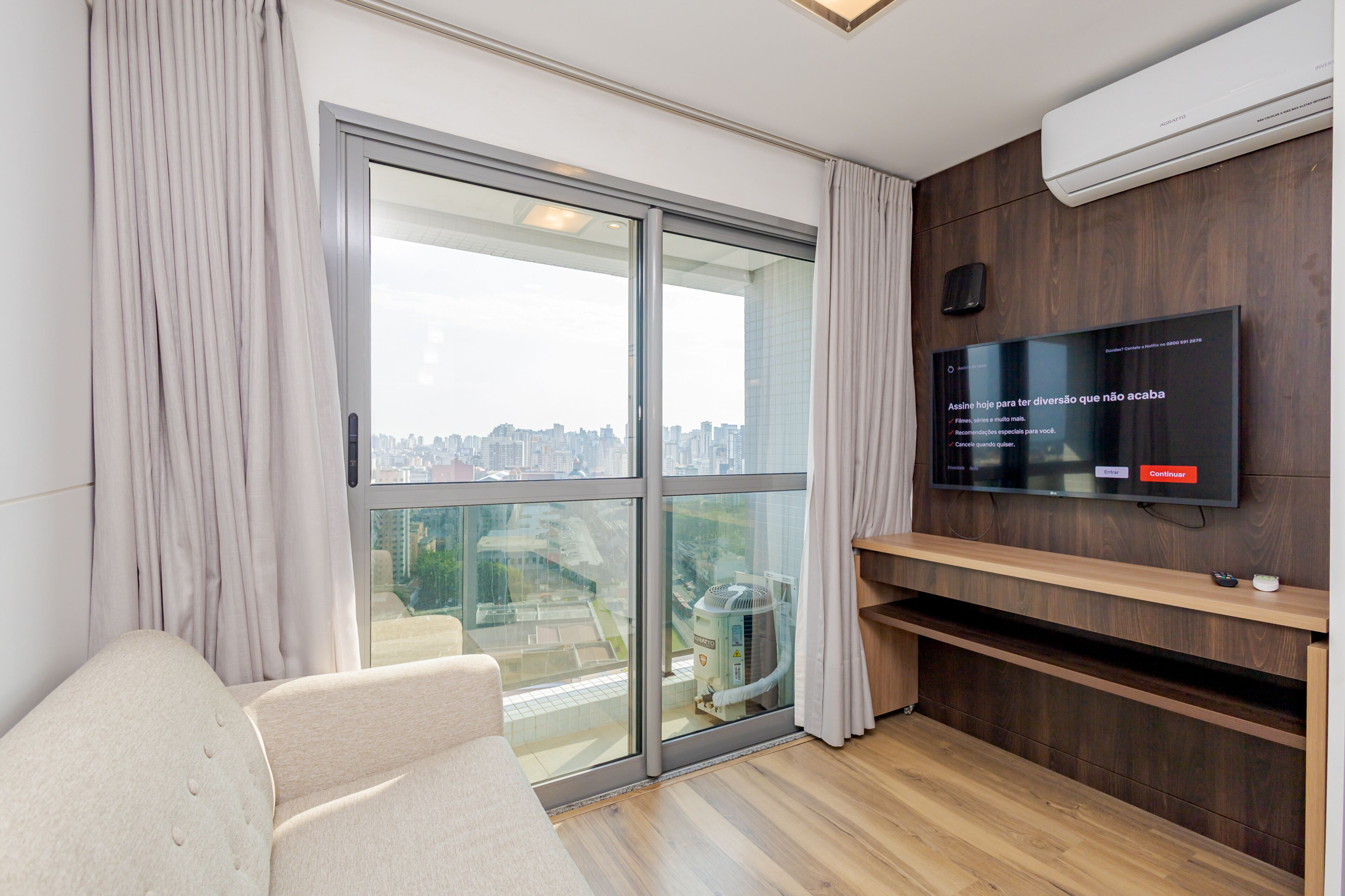 A modern living room with a large sliding glass door leading to a city view, a flat-screen TV mounted on a wooden wall, and a beige sofa.