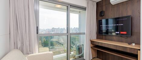 A modern living room with a large sliding glass door leading to a city view, a flat-screen TV mounted on a wooden wall, and a beige sofa.