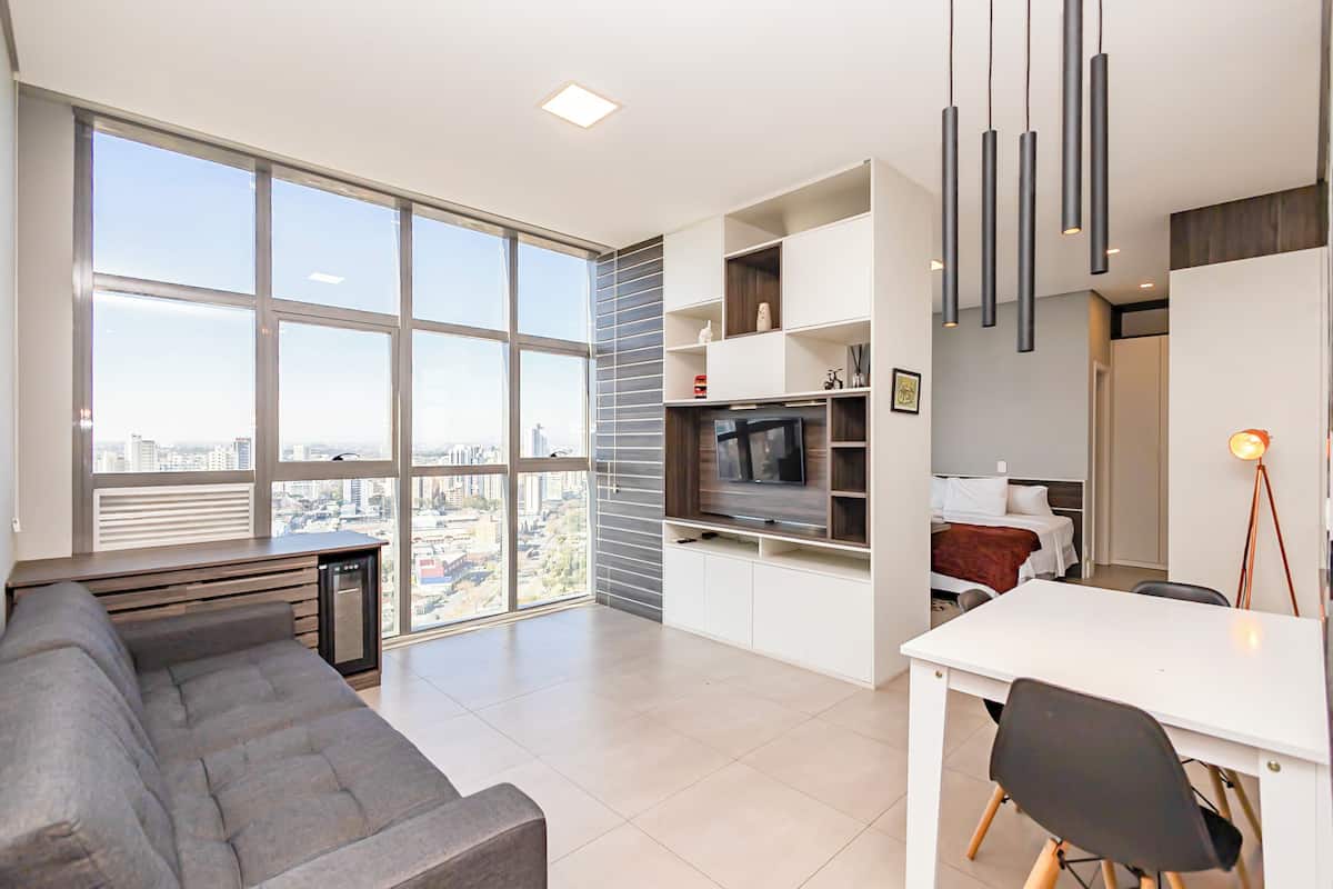 Sala de estar moderna com sofá, mesa de jantar e TV. Janelas amplas oferecem vista para a cidade.