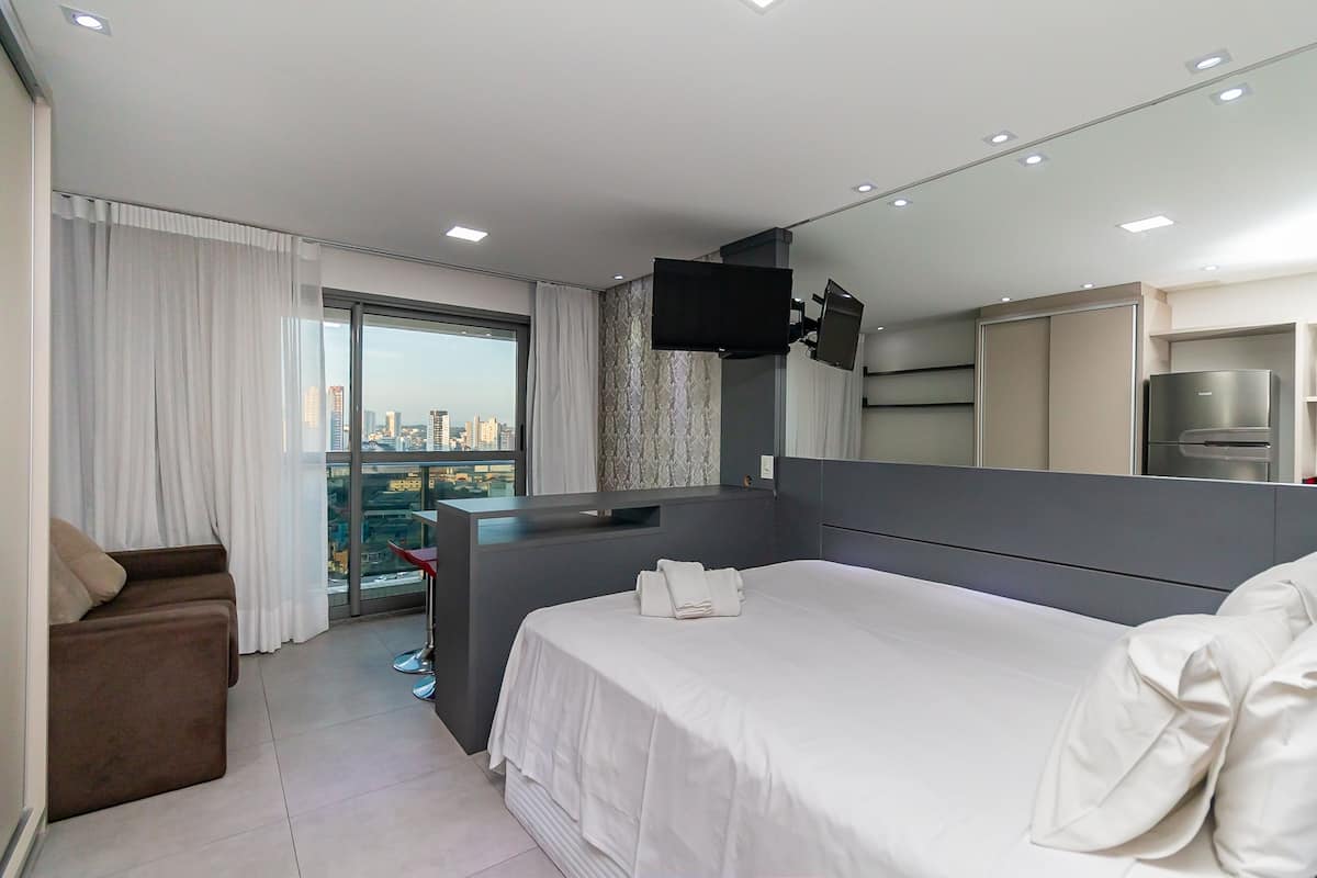 Quarto de hotel moderno com uma cama grande, área de estar, televisão e vista para a cidade.