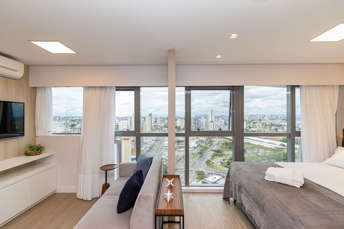 Quarto de hotel moderno com uma grande janela proporcionando vista para a cidade, uma cama, um sofá, uma mesa pequena e uma TV fixada na parede.