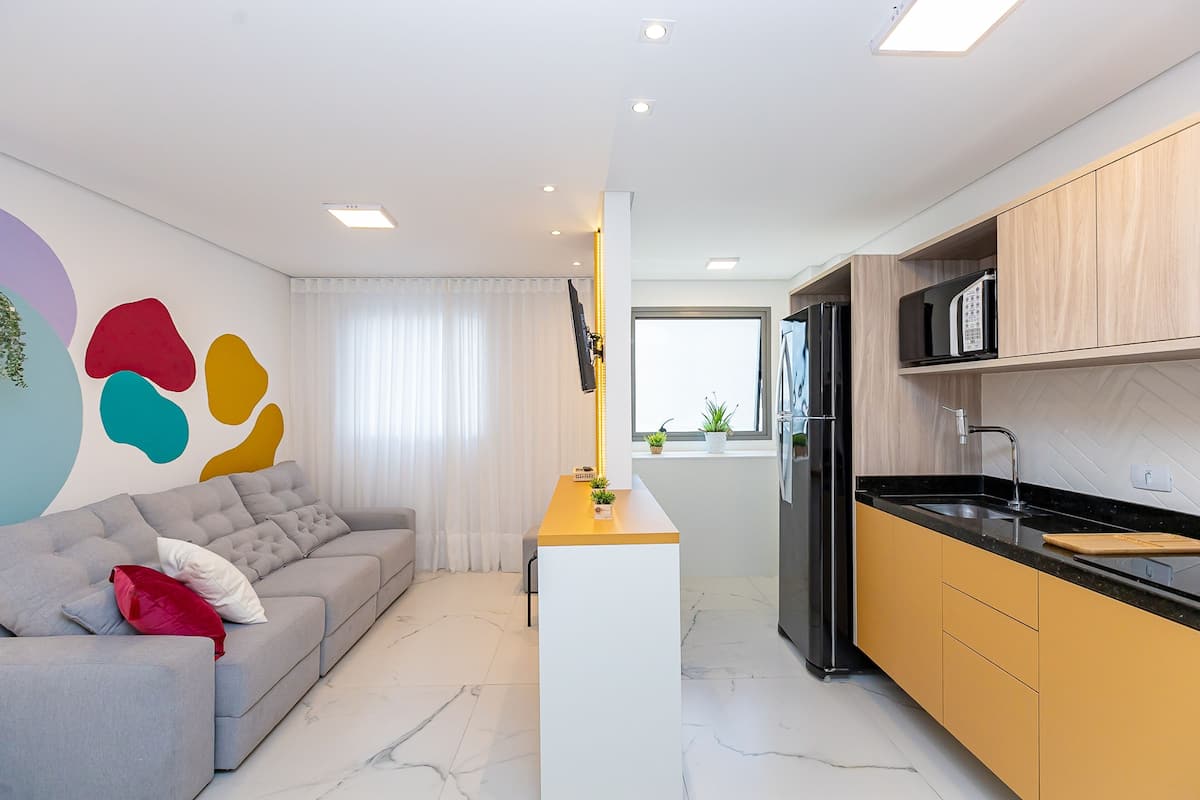 Uma sala moderna com um sofá cinzento, uma ilha pequena branca e uma cozinha com armários de madeira e bancada preta.