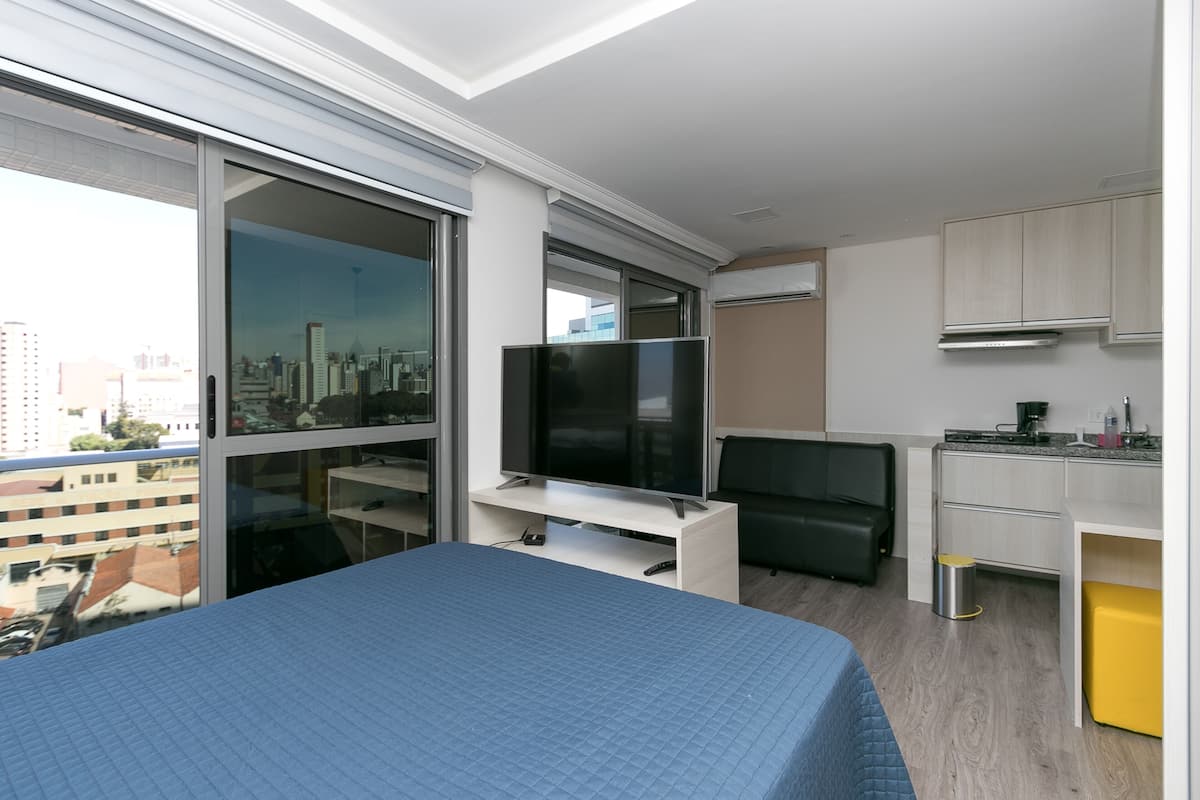 Quarto de hotel moderno com uma cama grande, televisão de tela plana, uma cozinha compacta e vista para a cidade através de grandes janelas.