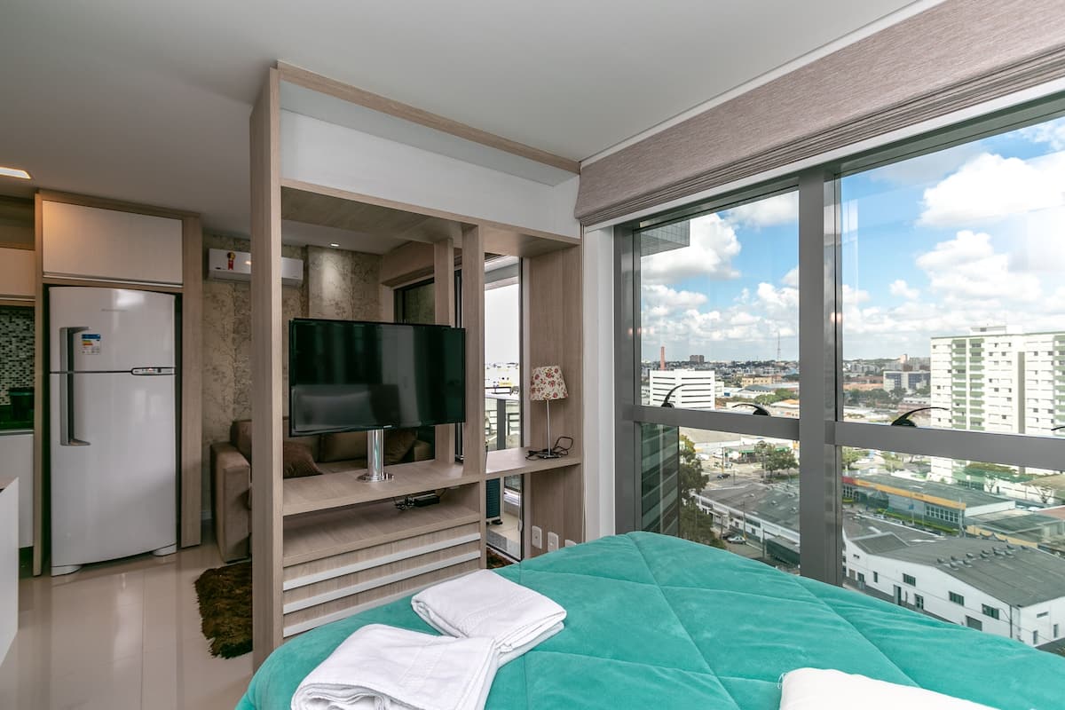 Quarto de hotel moderno com uma cama, televisão, frigorífico e uma grande janela com vista para a cidade.
