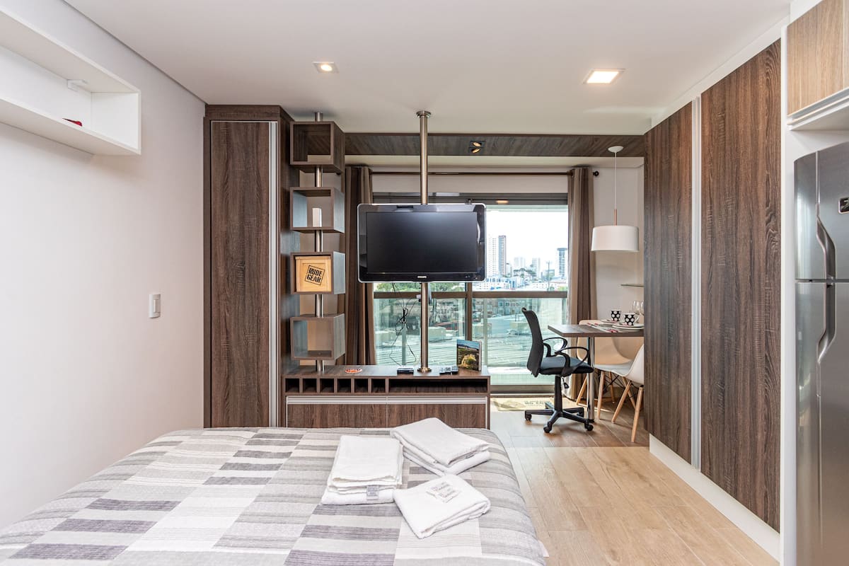 Quarto de hotel moderno com uma cama grande, televisão, escrivaninha com cadeira e vista para a cidade pela janela.