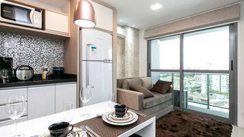 Cocina moderna equipada con microondas, refrigerador y cafetera. Se observa una mesa de comedor con un mantel individual y un tazón. En el fondo, un sofá con vista a la ciudad a través de amplios ventanales.