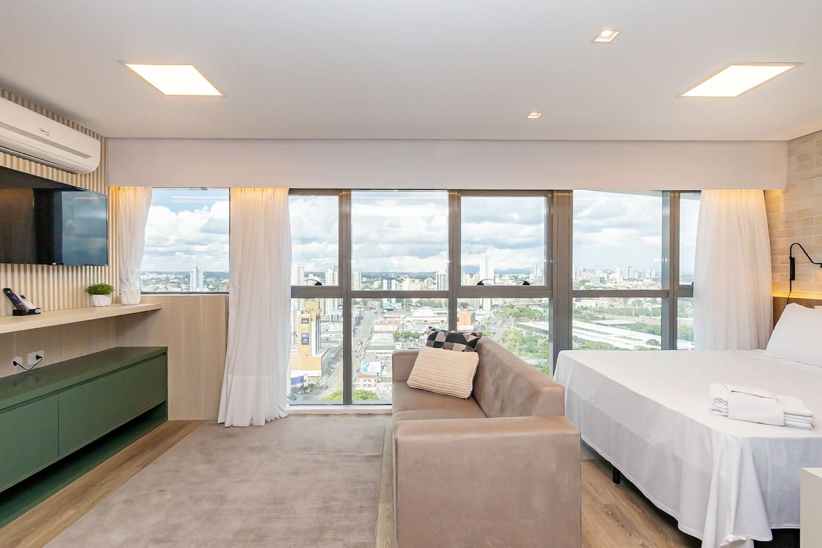 Quarto de hotel moderno com uma janela ampla, uma cama, um sofá, uma TV e um armário verde.