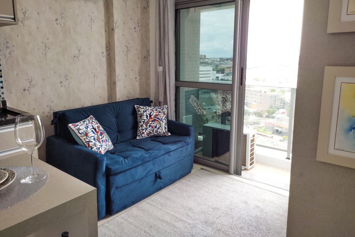 Uma sala de estar moderna com um sofá azul, duas almofadas estampadas, uma mesa de vidro com uma taça de vinho e vista para a cidade através de uma janela grande.