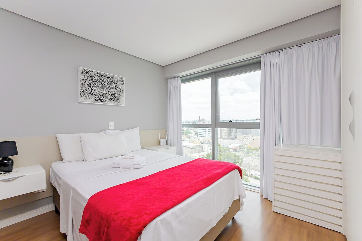 Quarto de hotel moderno com uma cama grande, mesa de cabeceira com abajur, janela com cortinas e vista para a cidade.