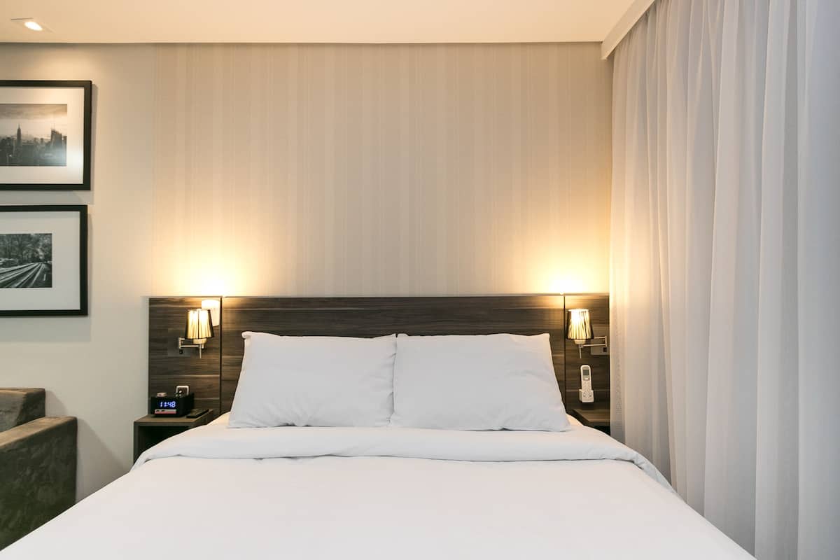 Quarto de hotel com cama bem arrumada, duas luminárias de cabeceira, mesa de cabeceira com telefone e relógio, e duas fotos emolduradas na parede.
