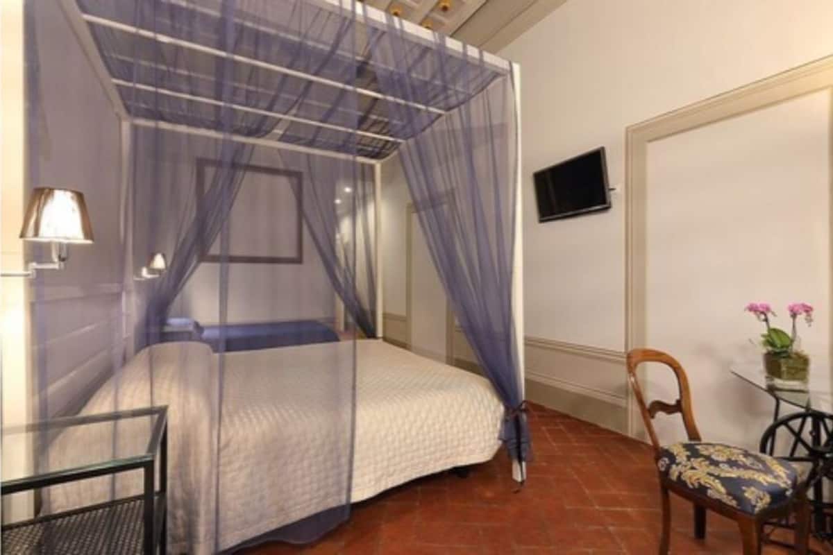 A bedroom with a bed, a chair, a table with a plant, and a TV on the wall.