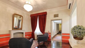 Um hall de hotel com duas poltronas, uma lareira, um espelho, cortinas vermelhas e uma planta sobre uma mesa de mármore.