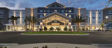 A hotel with a central entrance, multiple floors, and a landscaped front yard with palm trees.