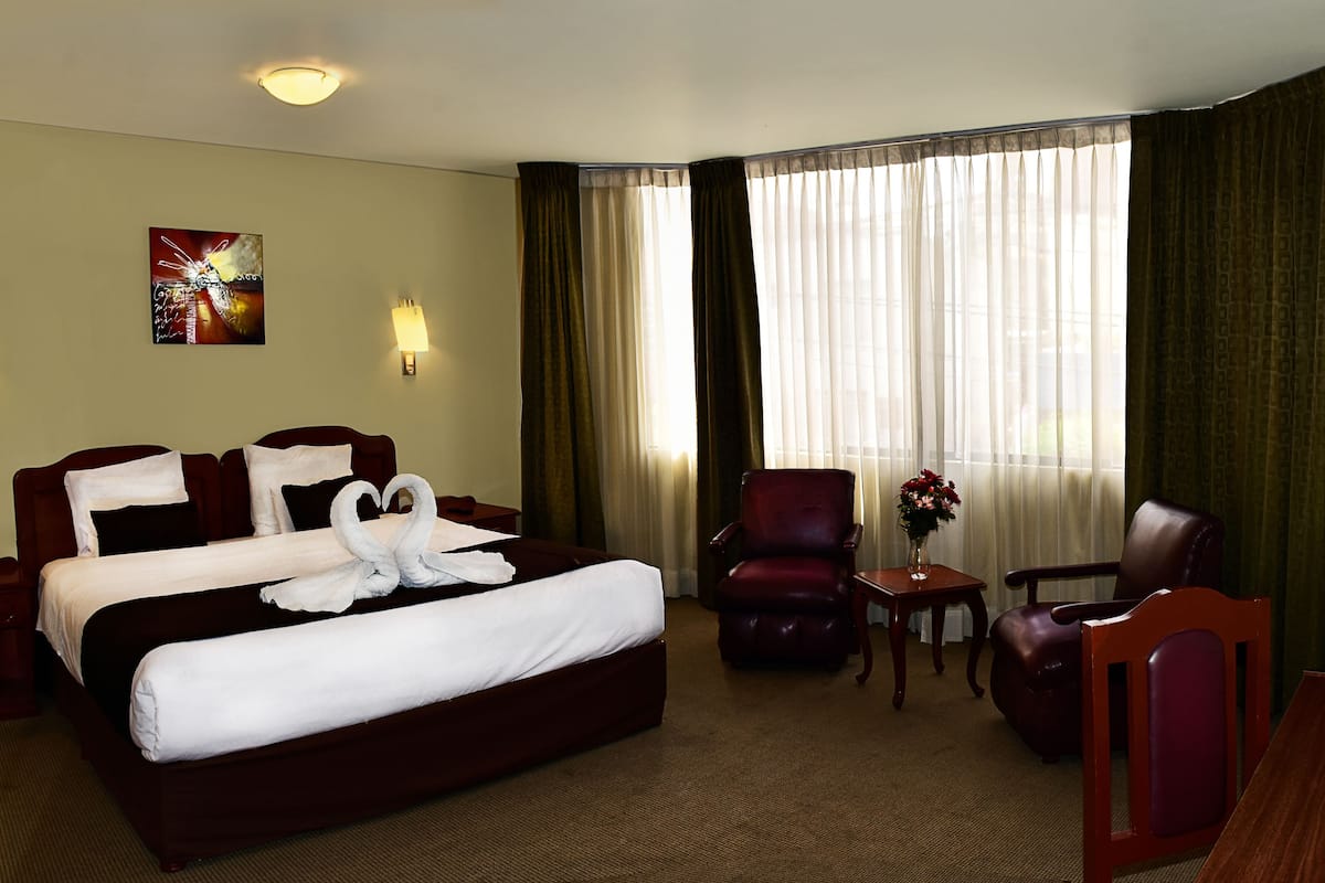 A hotel room with a large bed, two armchairs, a small table with a vase of flowers, a flat-screen TV mounted on the wall, and a painting on the wall.