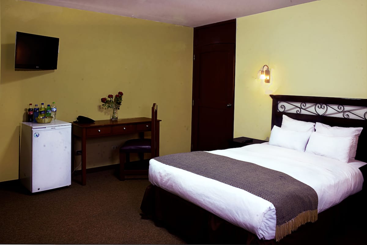 A hotel room with a bed, a desk, a refrigerator, a vase with flowers, and a wall-mounted TV.