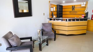 Un mostrador de recepción con un cartel de Riviera Hotels, dos sillones grises, una mesa de centro de cristal y un jarrón con plantas secas.