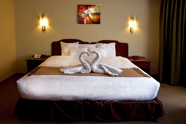 Una cama bien hecha con un arreglo de toallas en forma de corazón, un cuadro en la pared y dos lámparas fijadas a la pared.