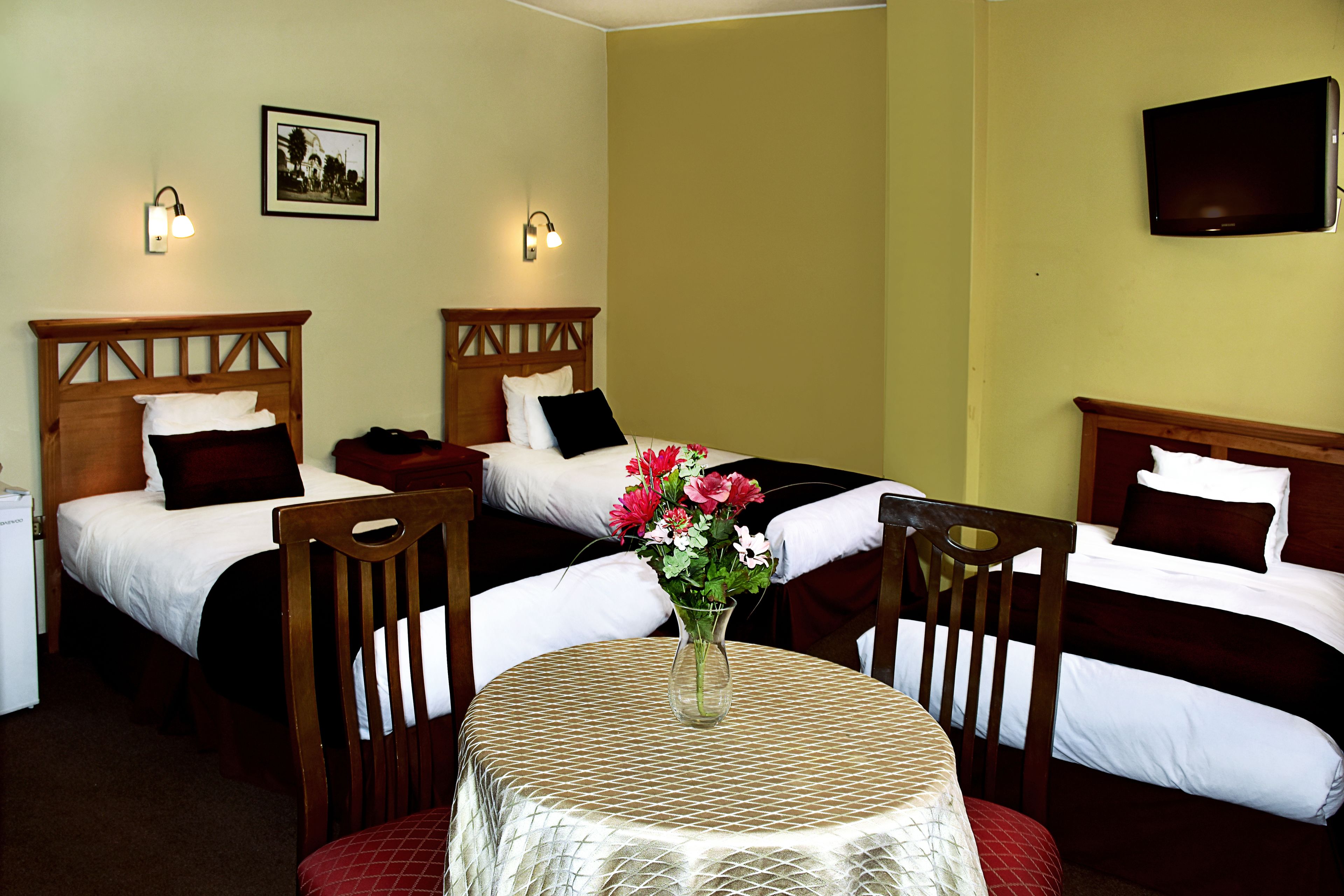 A hotel room with two single beds, a small table with a vase of flowers, and a television mounted on the wall.