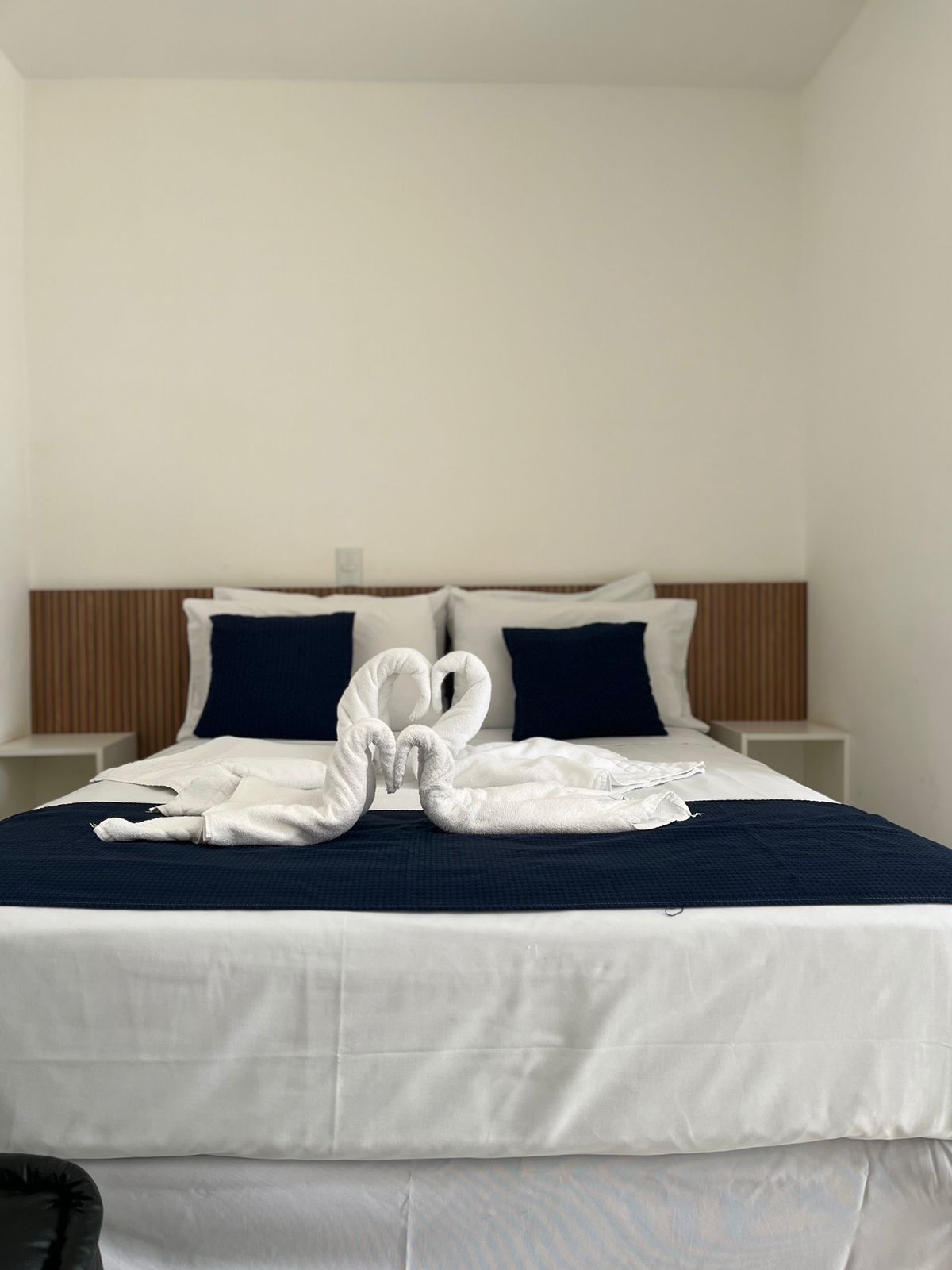 A neatly made bed with a white and navy blue color scheme, a wooden headboard, and a pair of towels arranged in a heart shape.