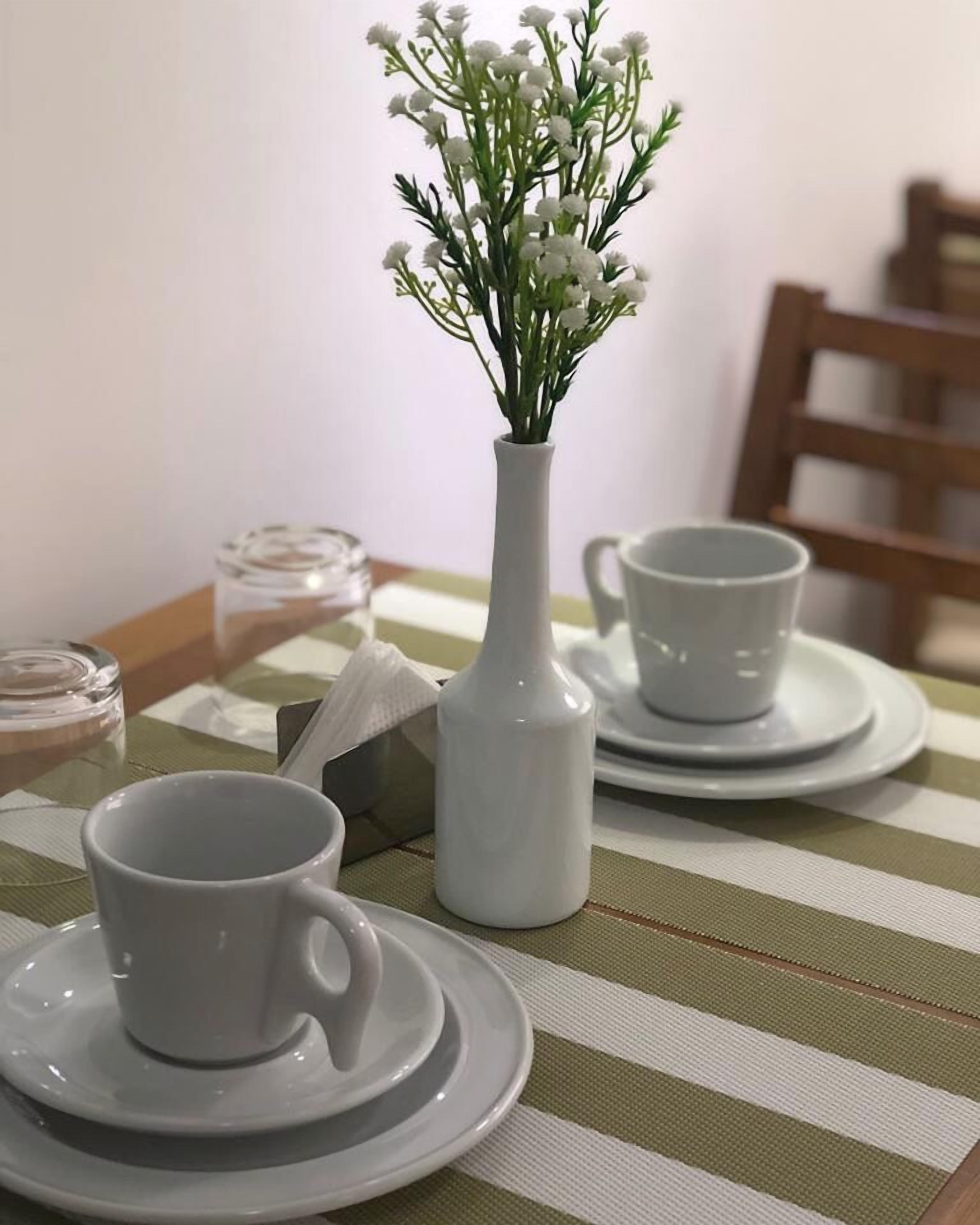 Una mesa de comedor con tazas y platillos blancos, un jarrón blanco con flores blancas y un mantel individual a rayas.