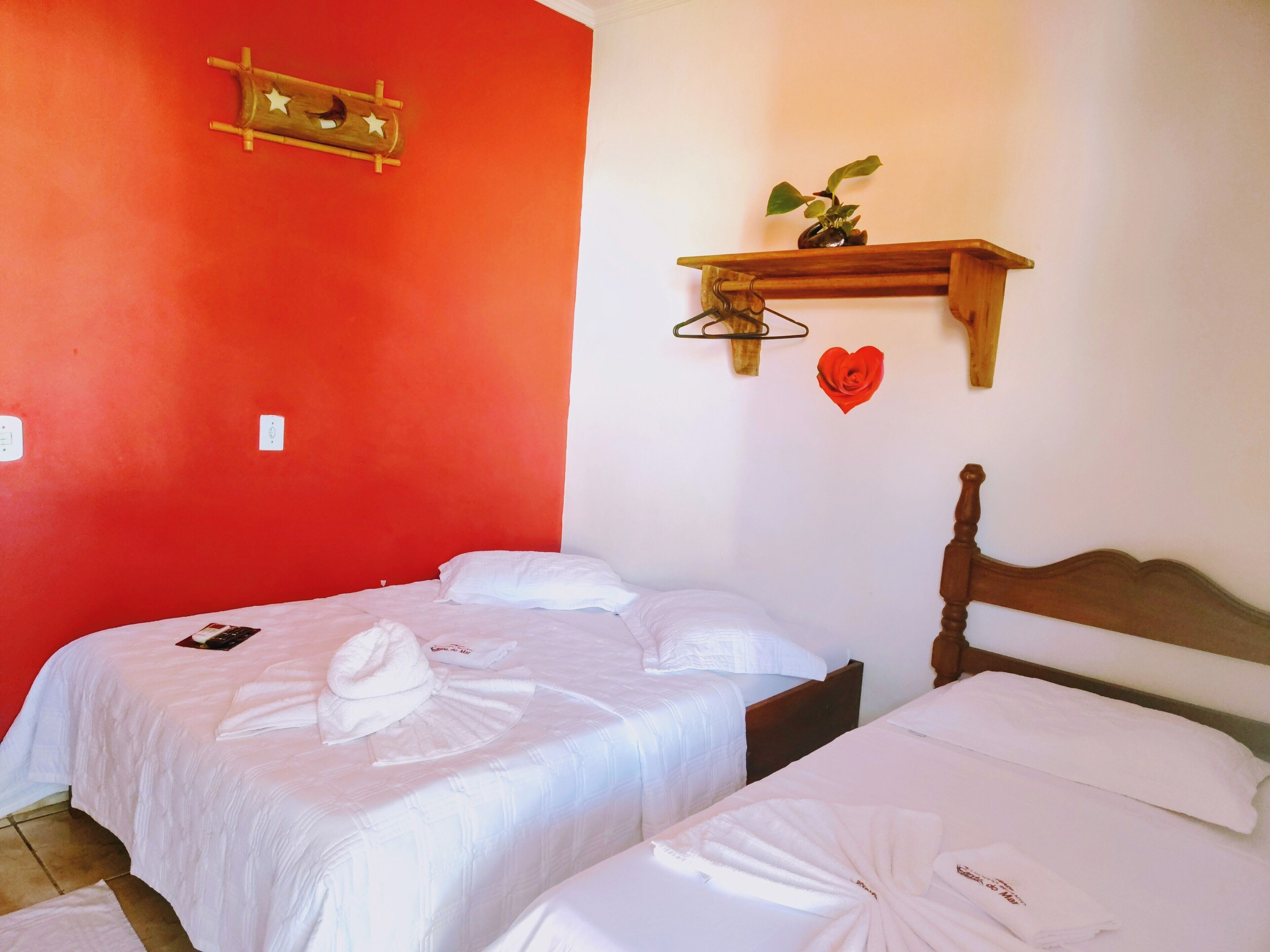 A hotel room with two beds, white bedding, a red accent wall, a wooden headboard, a small shelf with a plant and a heart decoration, and a wall-mounted hook.