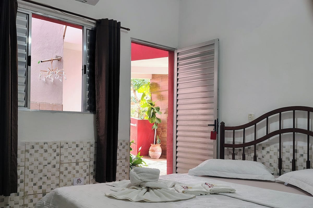 Quarto com cama, janela com cortinas, aparelho de ar condicionado e vista para o exterior.