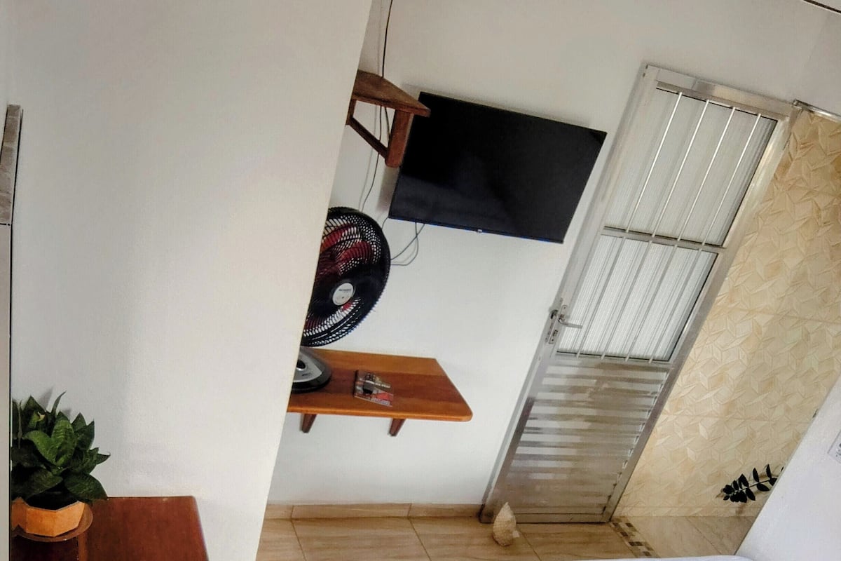 Quarto compacto com cama, uma pequena estante de madeira, uma planta em vaso e um ventilador.