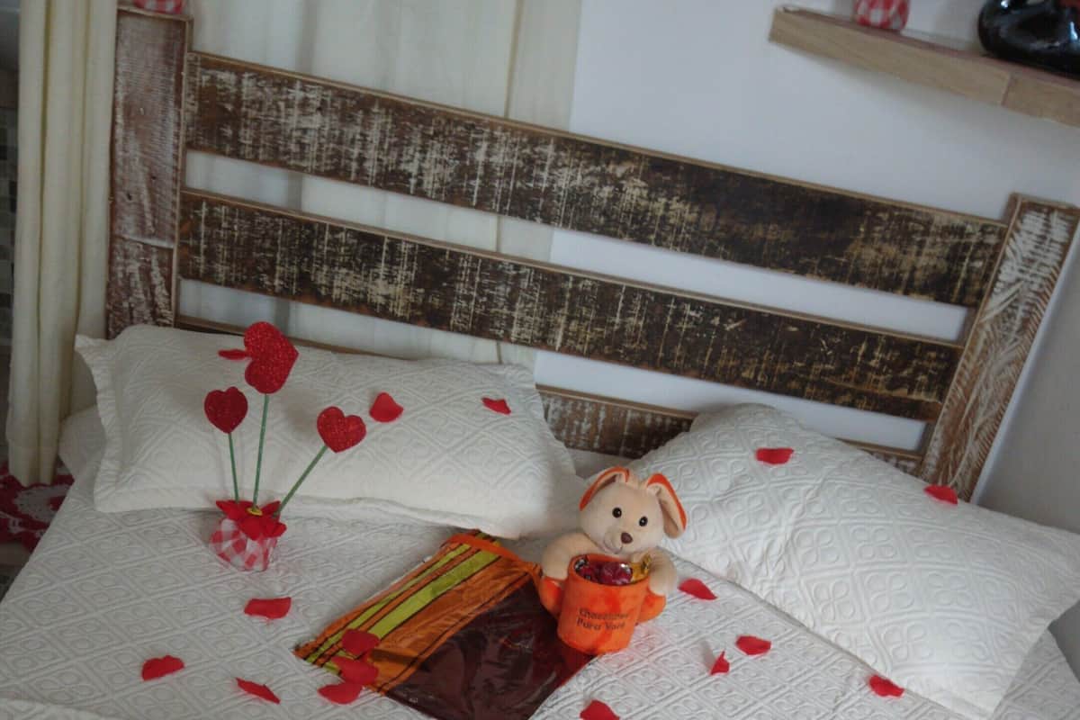 Uma cama com um ursinho de pelúcia, um pequeno vaso com flores vermelhas e uma cabeceira de madeira.
