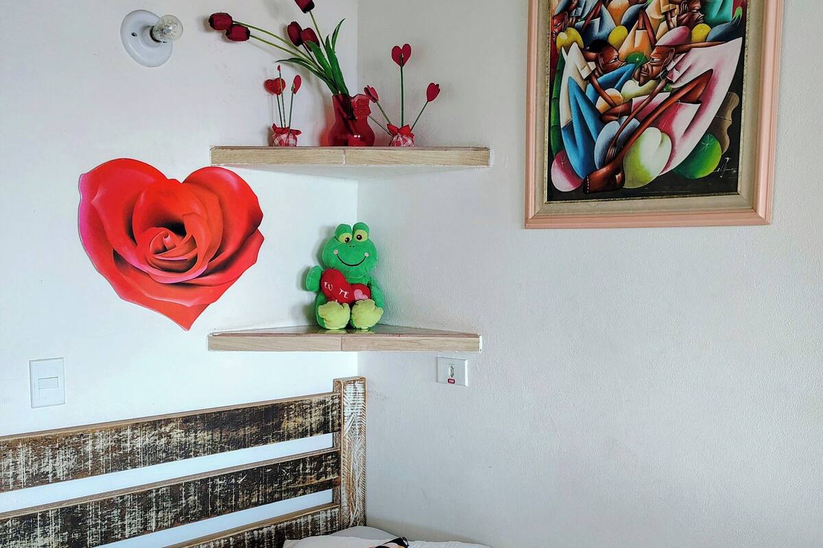 Quarto com cabeceira de madeira, uma grande decoração de flor vermelha, um brinquedo de pelúcia em formato de sapo verde e uma pintura abstrata emoldurada.
