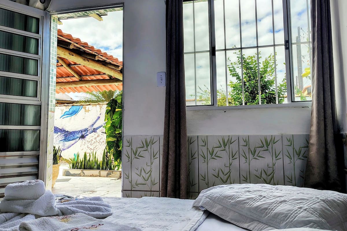 Quarto de hotel com cama, janela com cortinas, aparelho de ar condicionado e vista para o exterior.