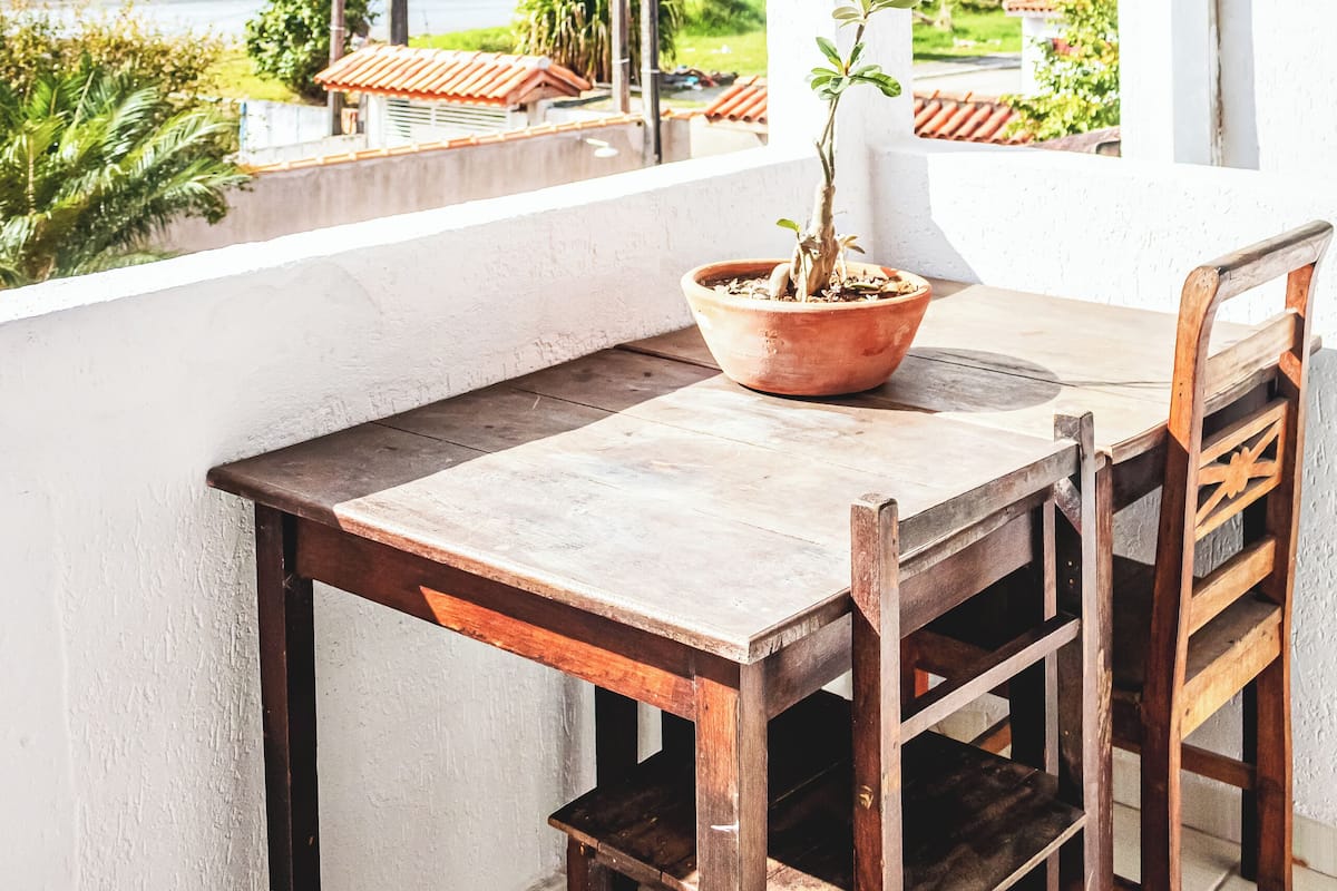 Varanda com mesa e cadeiras de madeira, um vaso de planta e vista para o mar e montanhas.