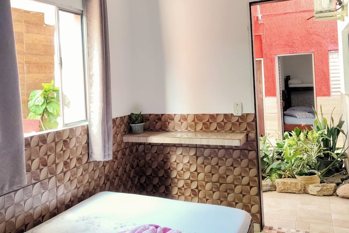 Um quarto com cama, janela com cortinas, ventilador de teto, pequena prateleira com uma planta e vista de um prédio vermelho lá fora.