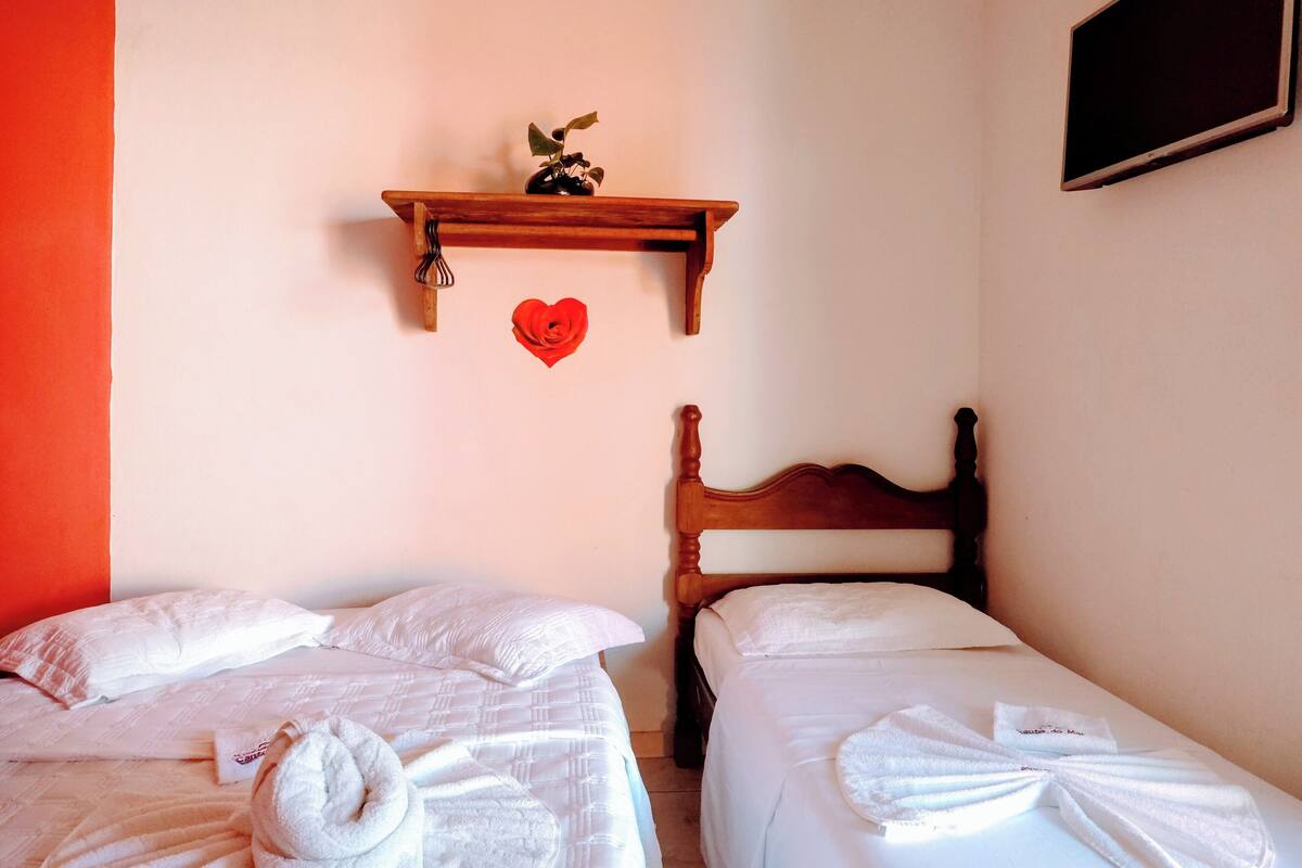 Quarto com duas camas, cabeceira de madeira, prateleira com decoração de coração e televisão fixada na parede.